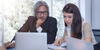 an older man and a woman sitting at a table looking at a laptop